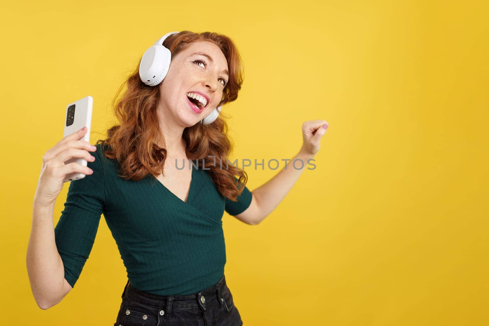 Cool redheaded woman listening to music with headphones and mobile in studio with yellow background