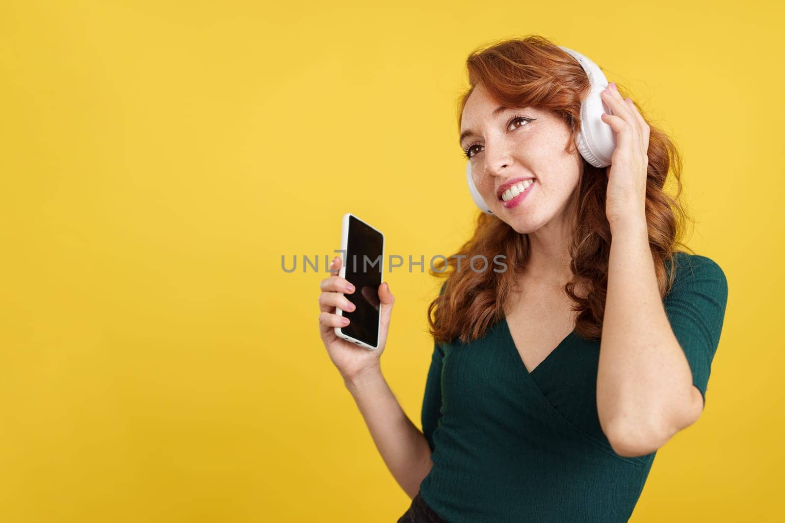 Relaxed redheaded woman listening to music looking up in studio with yellow background