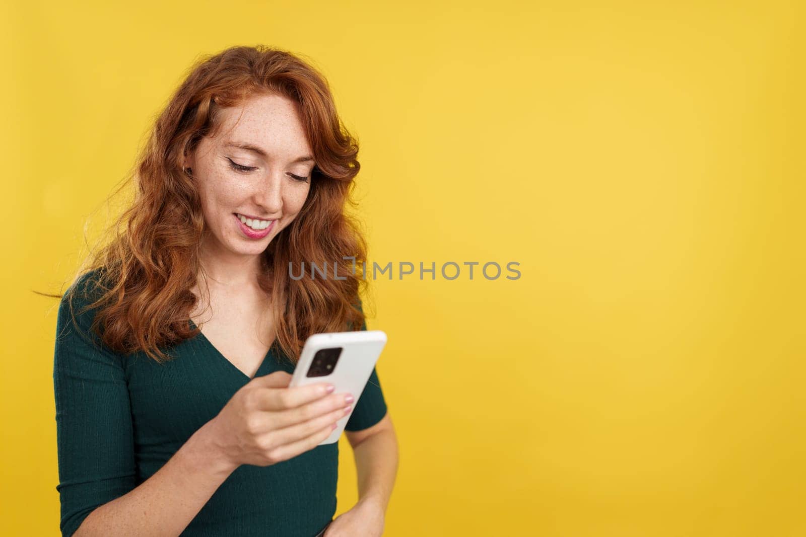 Happy redheaded woman smiling while using the mobile in studio with yellow background