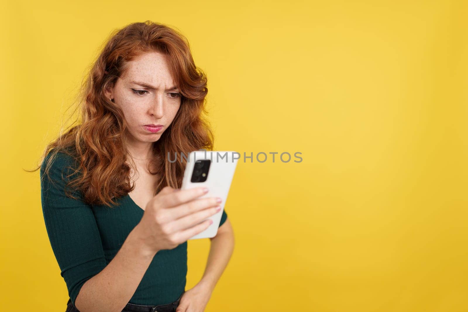 Worried redheaded woman using a mobile phone in studio with yellow background