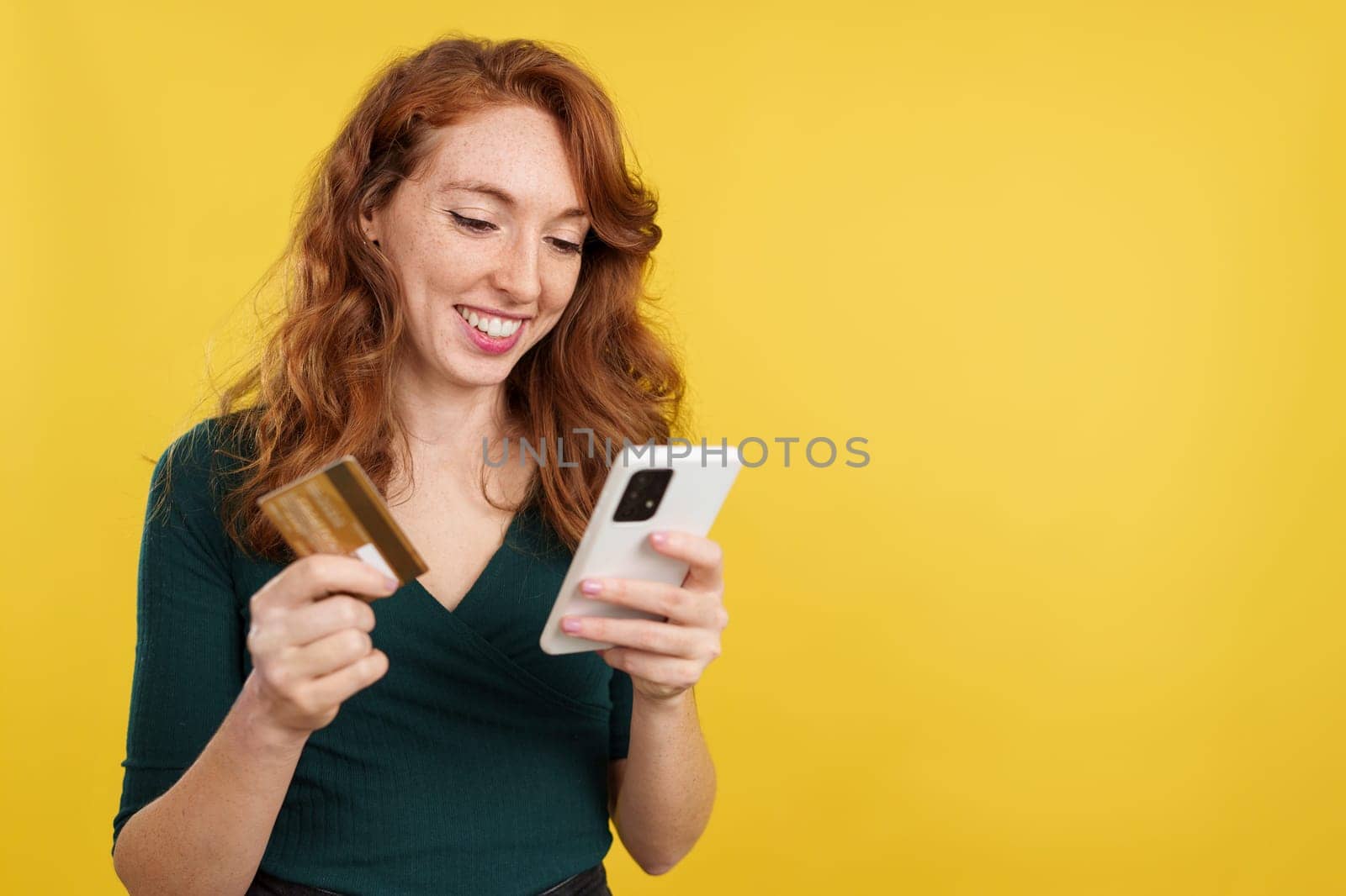 Happy redheaded woman shopping online with the mobile and a card in studio with yellow background