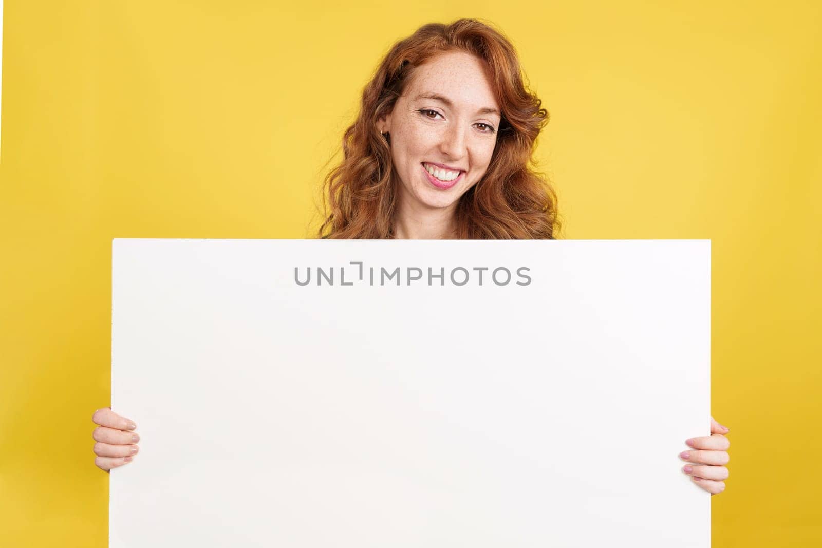 Redheaded woman holding a blank panel while smiling at the camera in studio with yellow background