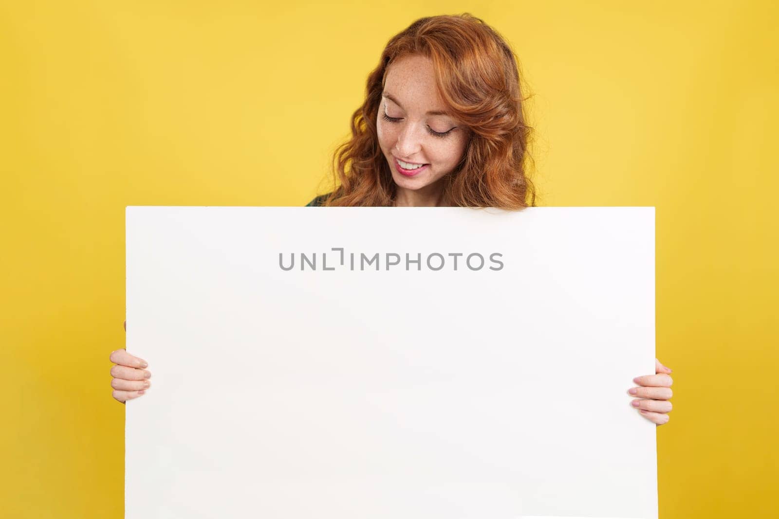 Redheaded woman looking down to the panel she held in studio with yellow background