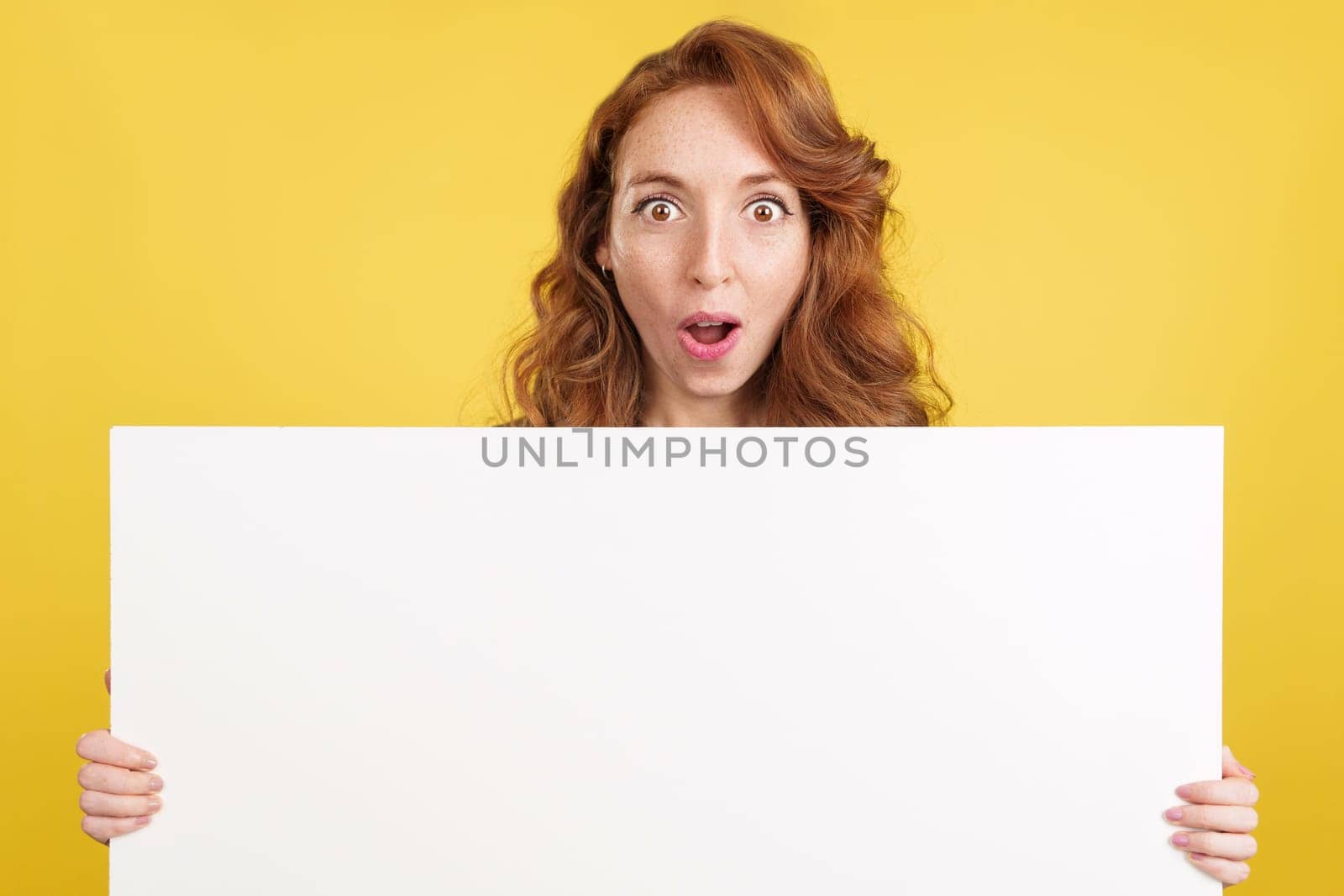 Amazed redheaded woman holding a blank panel while looking at camera in studio with yellow background