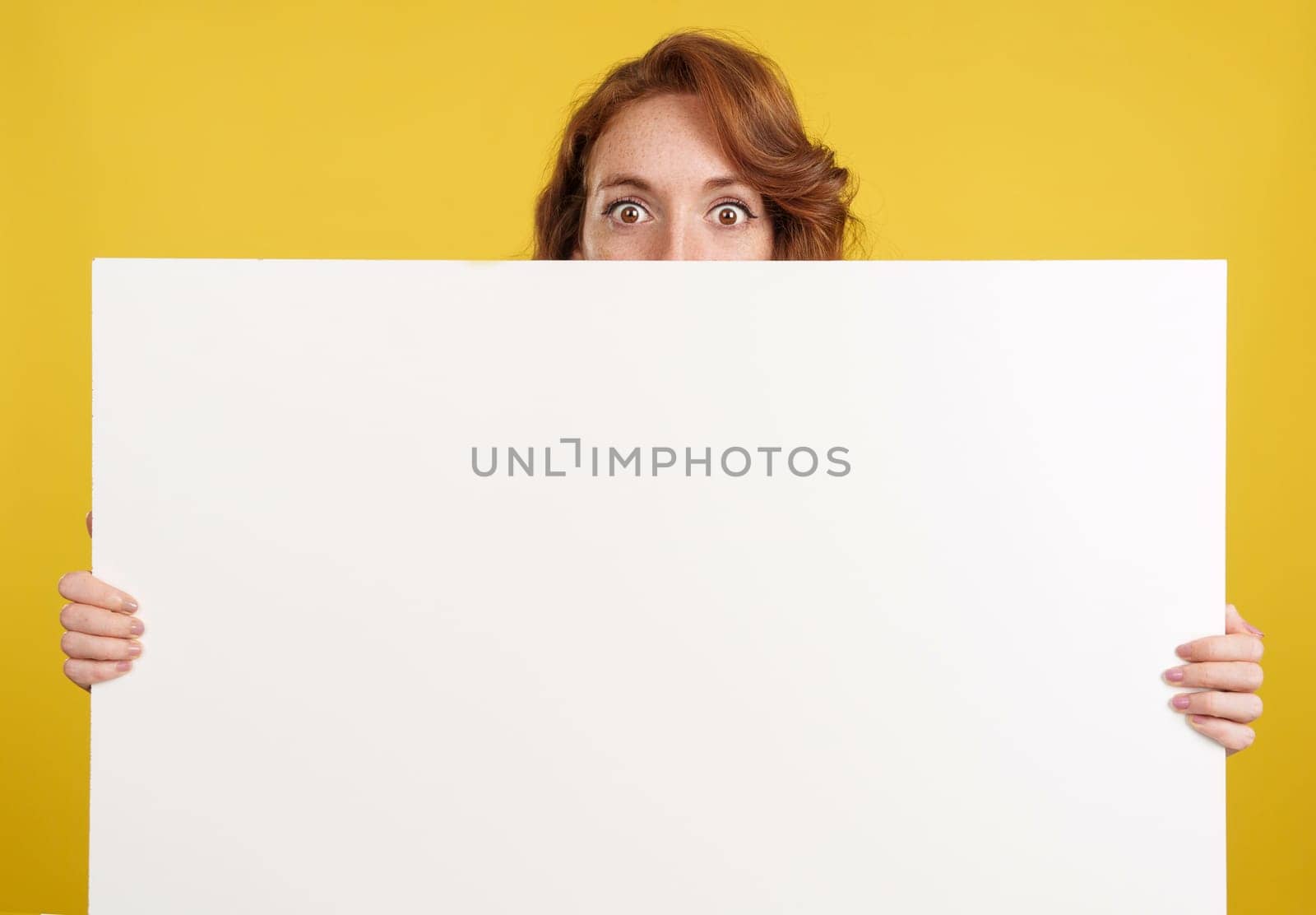 Surprised redheaded woman hiding behind a blank panel in studio with yellow background