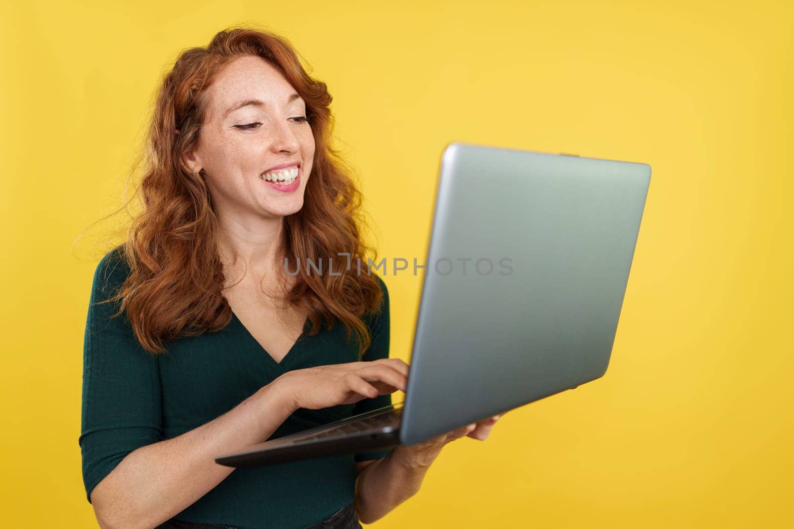 Happy redheaded woman using a laptop while smiling in studio with yellow background