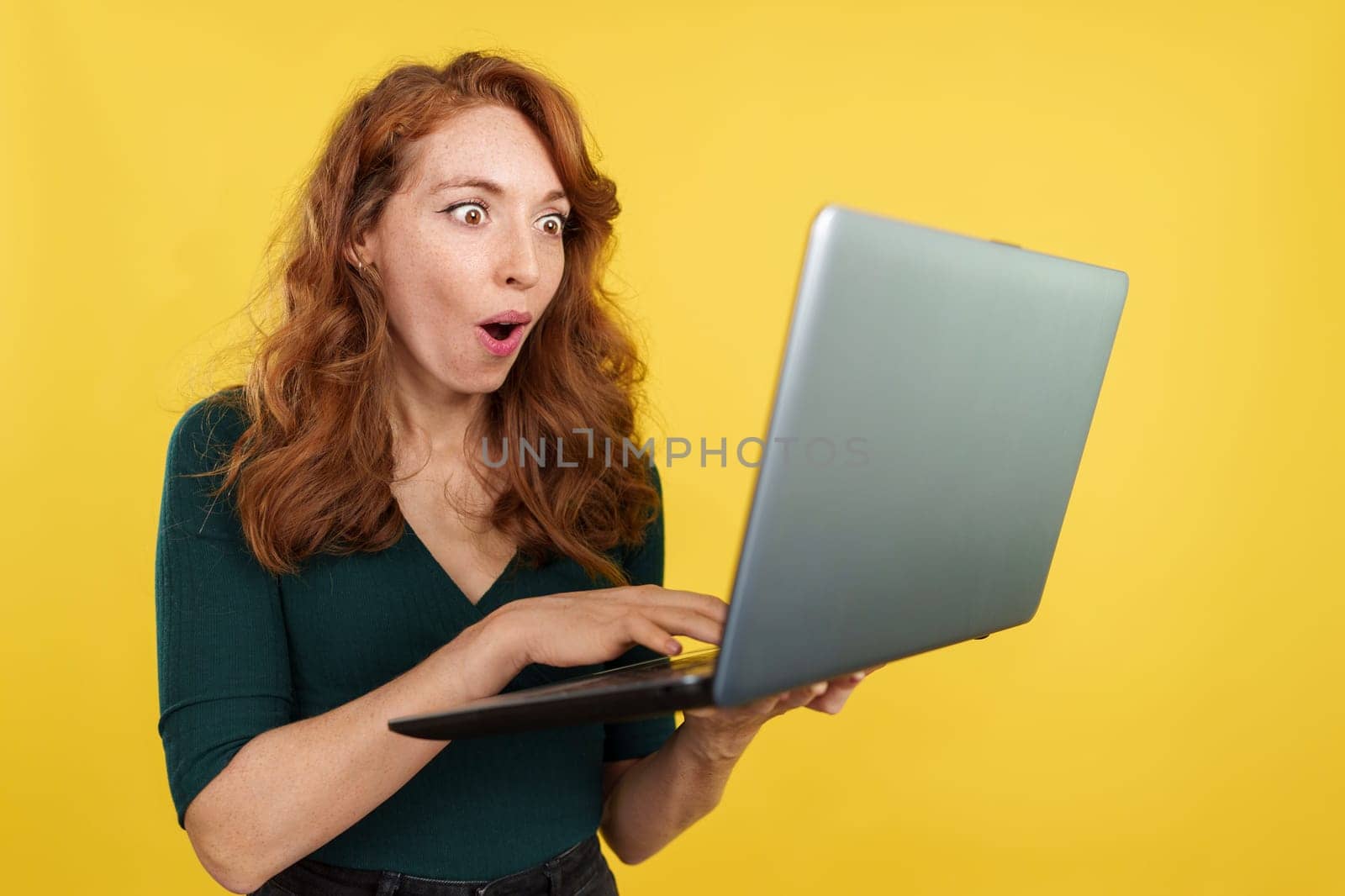Surprised redheaded woman using a laptop while smiling in studio with yellow background
