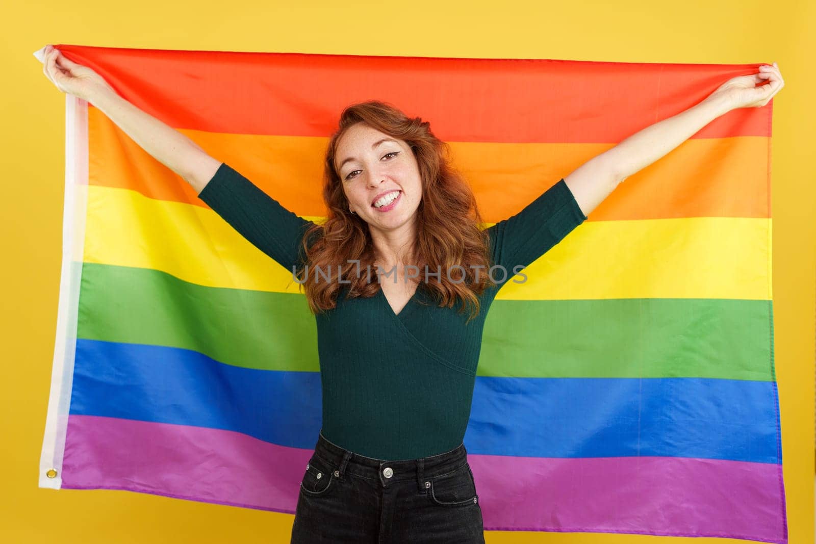 Happy redheaded woman raising a lgbt rainbow flag in studio with yellow background