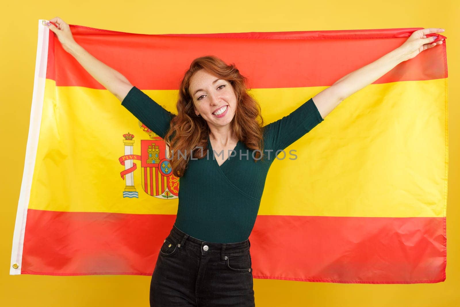 Happy redheaded woman smiling and raising a spanish national flag in studio with yellow background