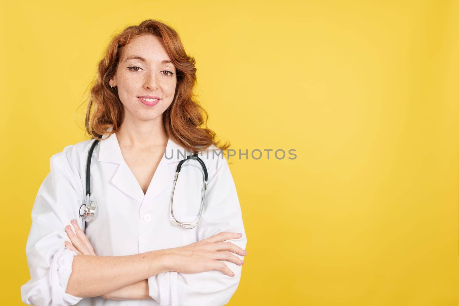 Redheaded female doctor smiling at the camera with arms crossed in studio with yellow background