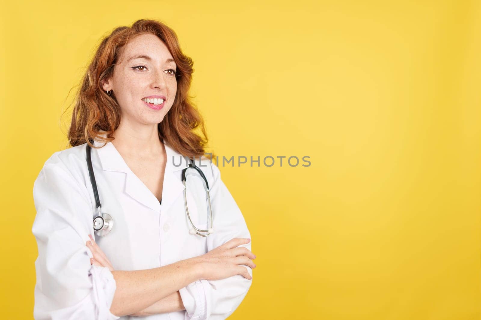 Friendly redheaded female doctor in uniform and stethoscope in studio with yellow background