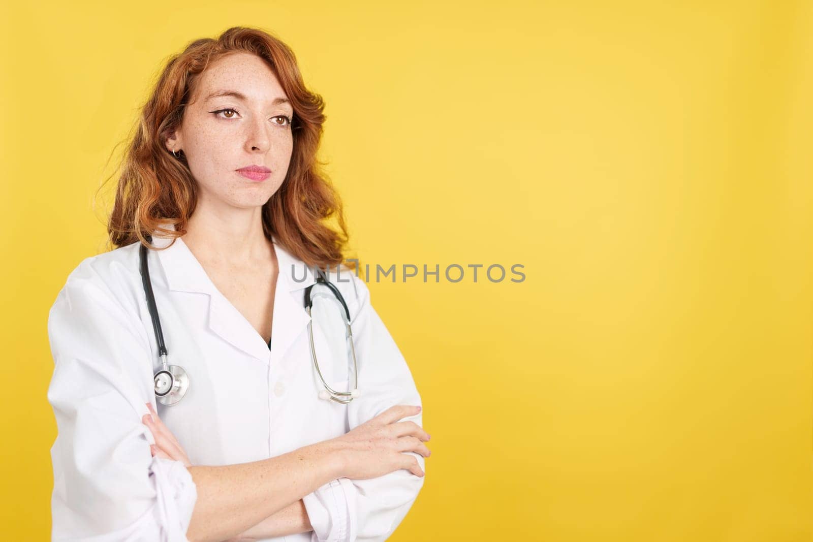 Serious redheaded female doctor with arms crossed looking away in studio with yellow background