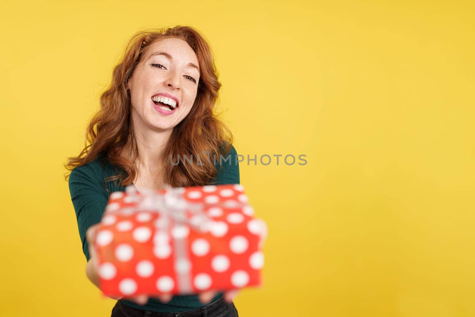 Happy redheaded woman looking at the camera giving a present in studio with yellow background