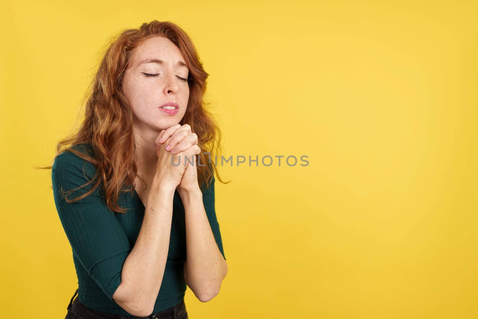 Redheaded woman praying with folded hands and eyes closed in studio with yellow background