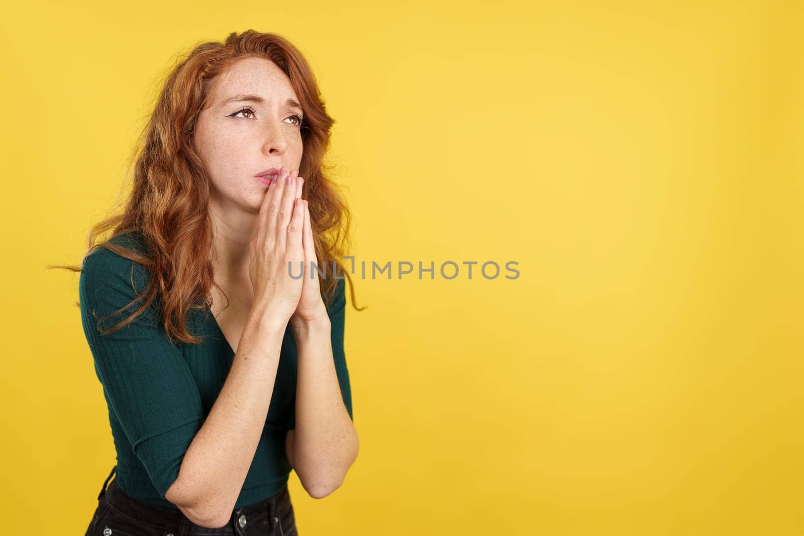 Redheaded woman looking up while praying with folded hands in studio with yellow background