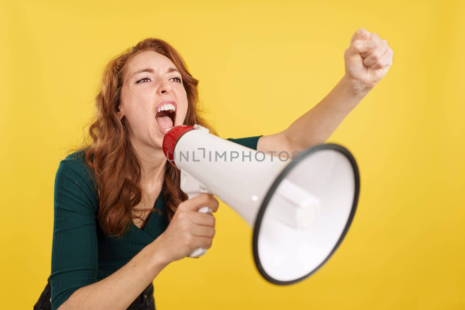 Upset redheaded woman yelling using a loudspeaker in studio with yellow background