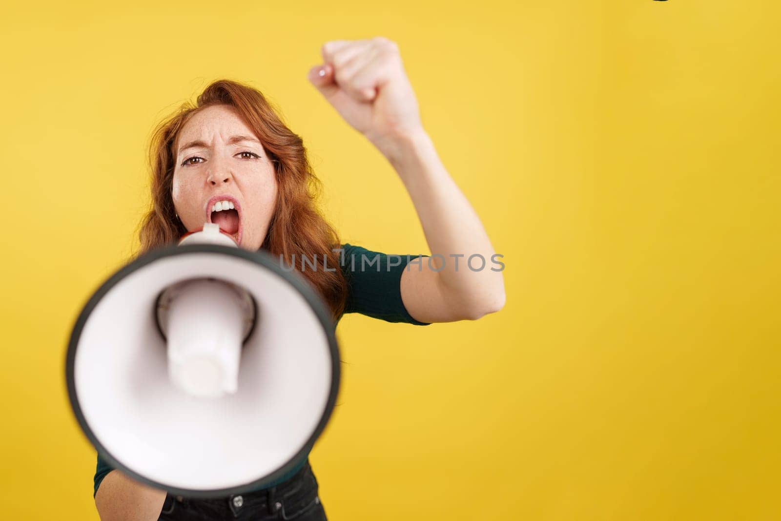 Angry redheaded woman shouting using a loudspeaker in studio with yellow background