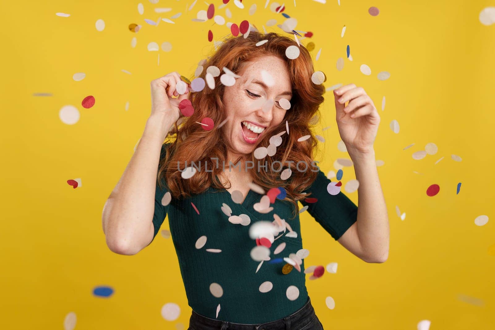 Happy redheaded woman dancing surrounded by confetti flying in the air in studio with yellow background