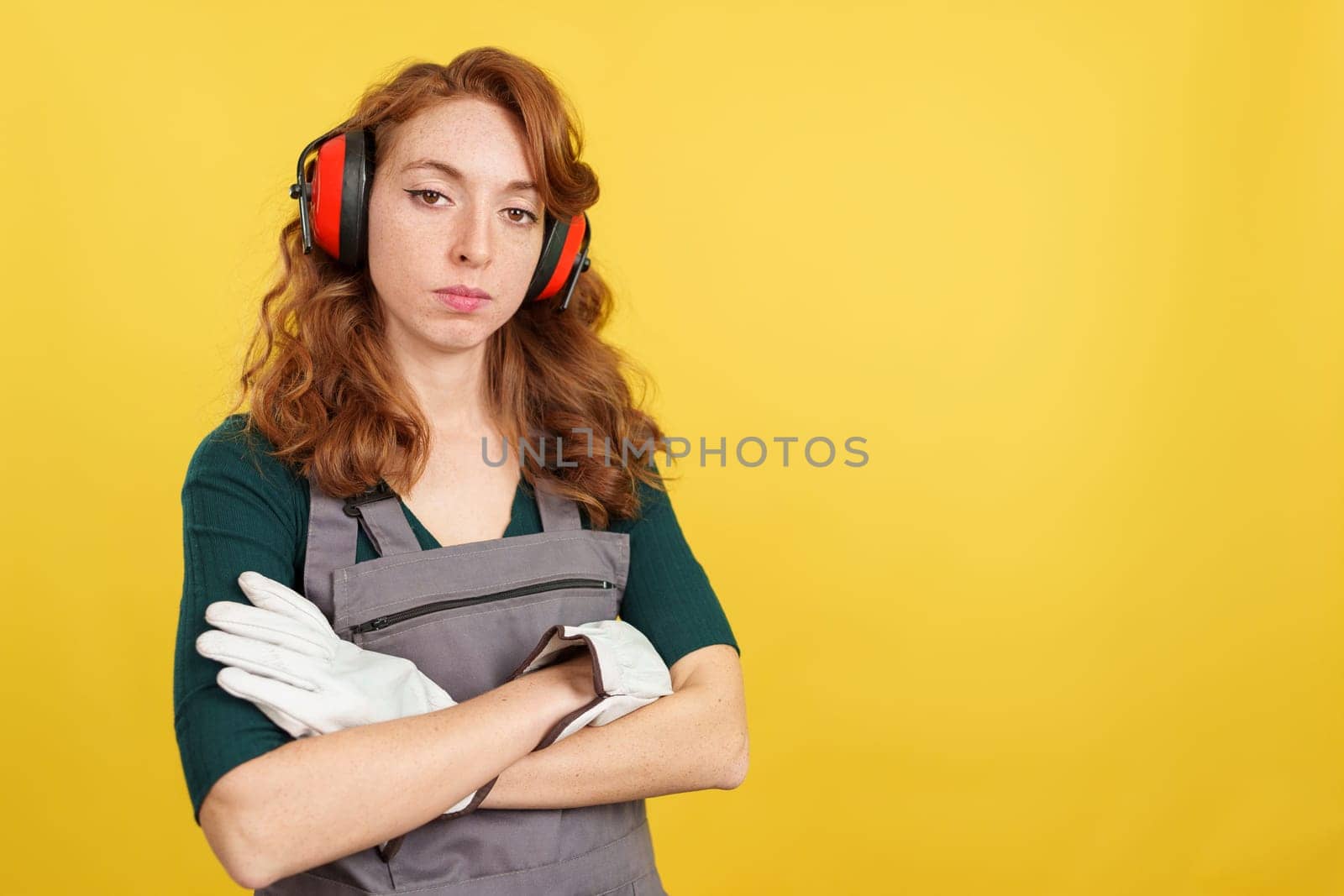 Serious redheaded female worker standing with arms crossed looking at camera in studio with yellow background