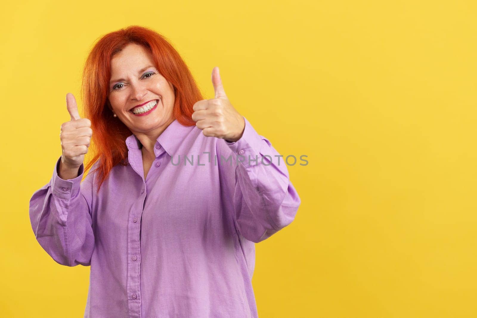 Happy mature woman gesturing agreement raising a thumb up in studio with yellow background