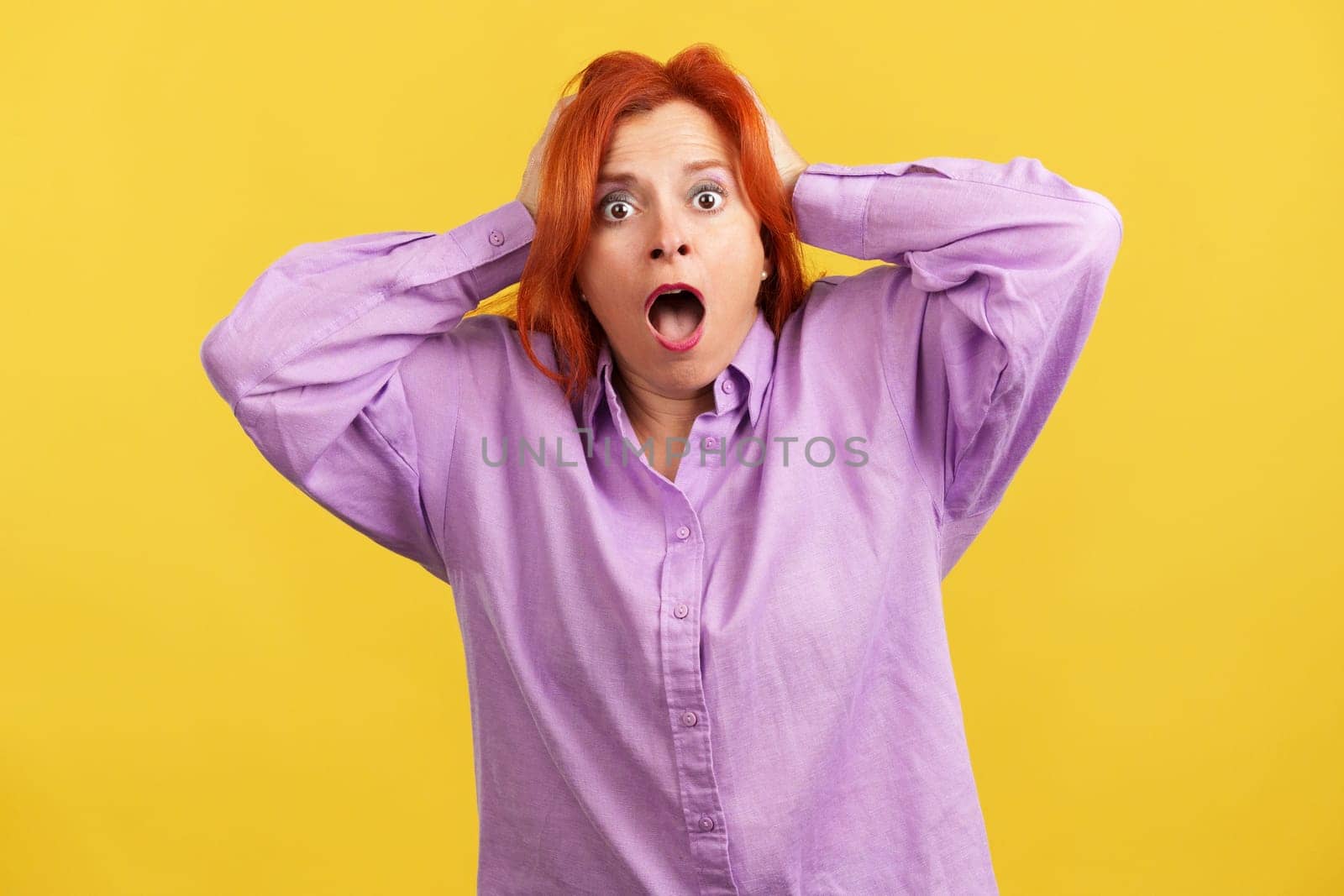 Surprised mature woman with red hair dye gesturing and looking at the camera in studio with yellow background