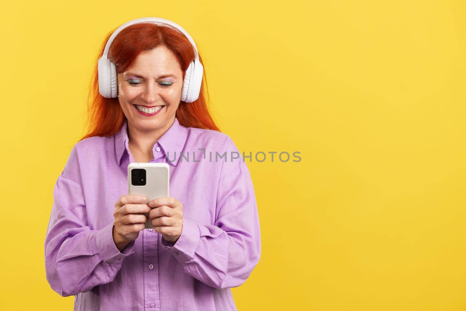 Mature woman listening to music with headphones and a mobile in studio with yellow background