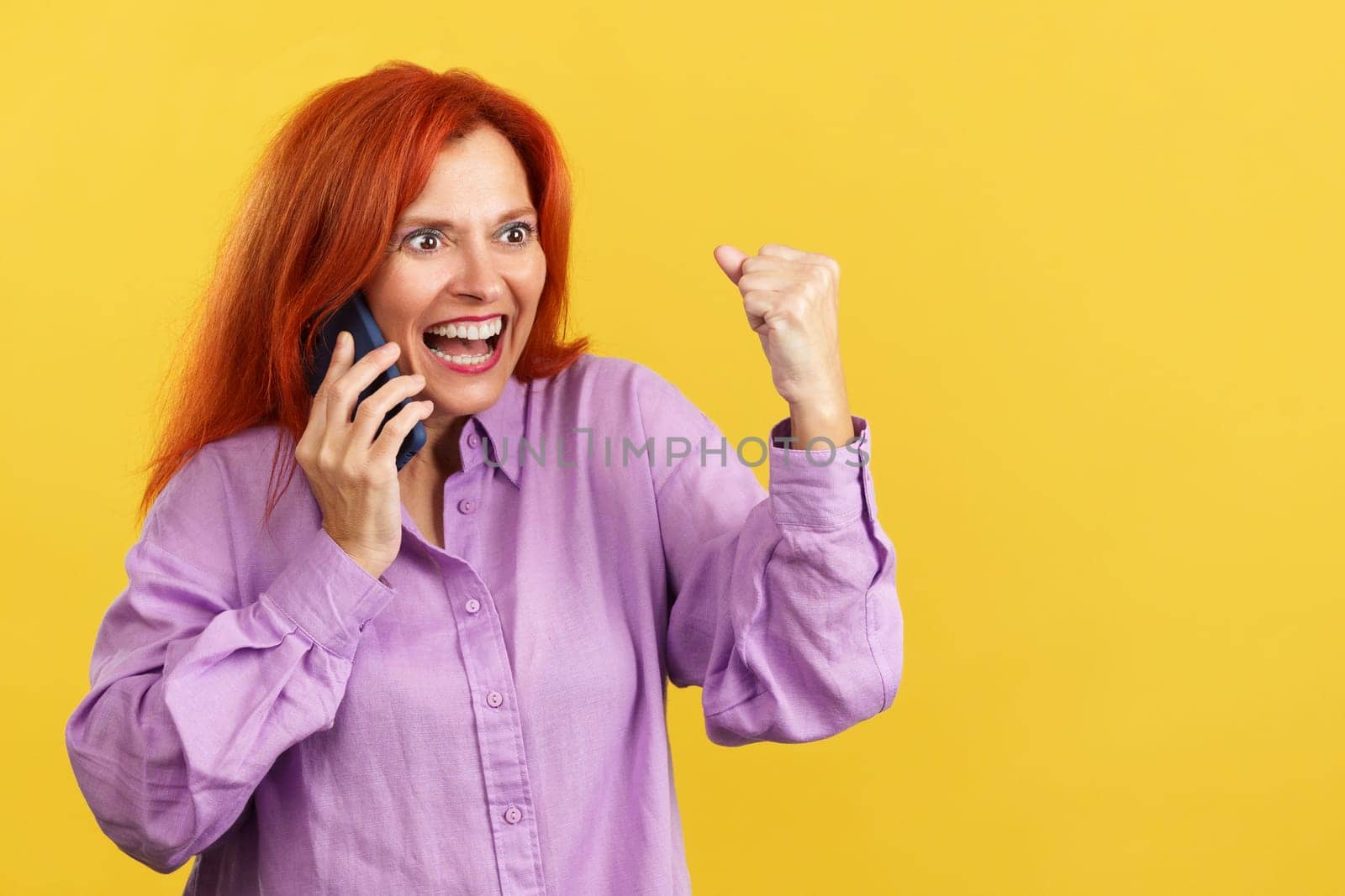 Happy mature redheaded woman celebrating while taking to the mobile in studio with yellow background