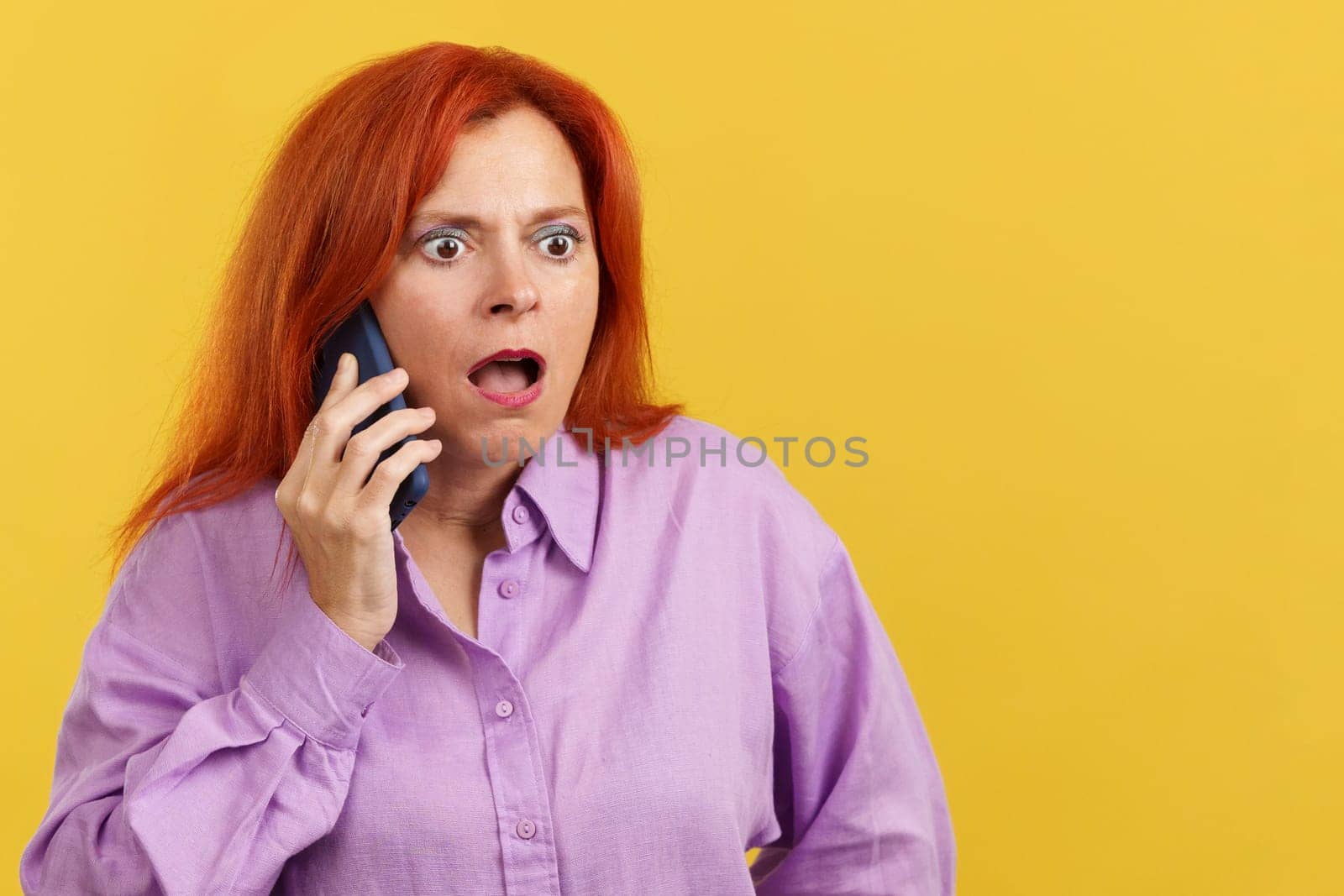 Surprised mature redheaded woman talking to the mobile in studio with yellow background