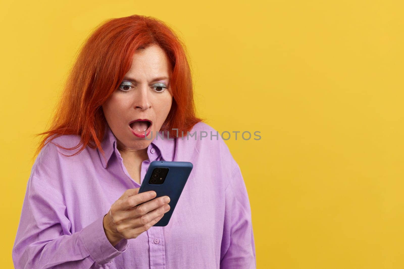 Surprised redheaded mature woman looking the screen of a mobile in studio with yellow background