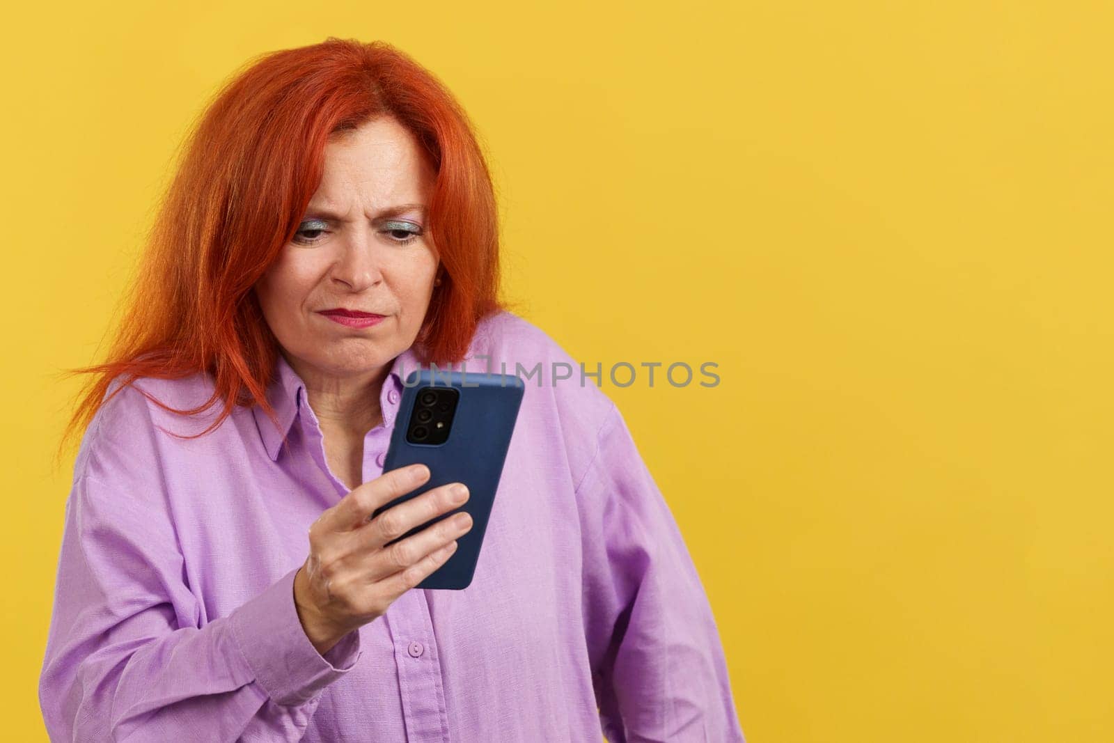 Worried redheaded mature woman using a mobile phone in studio with yellow background