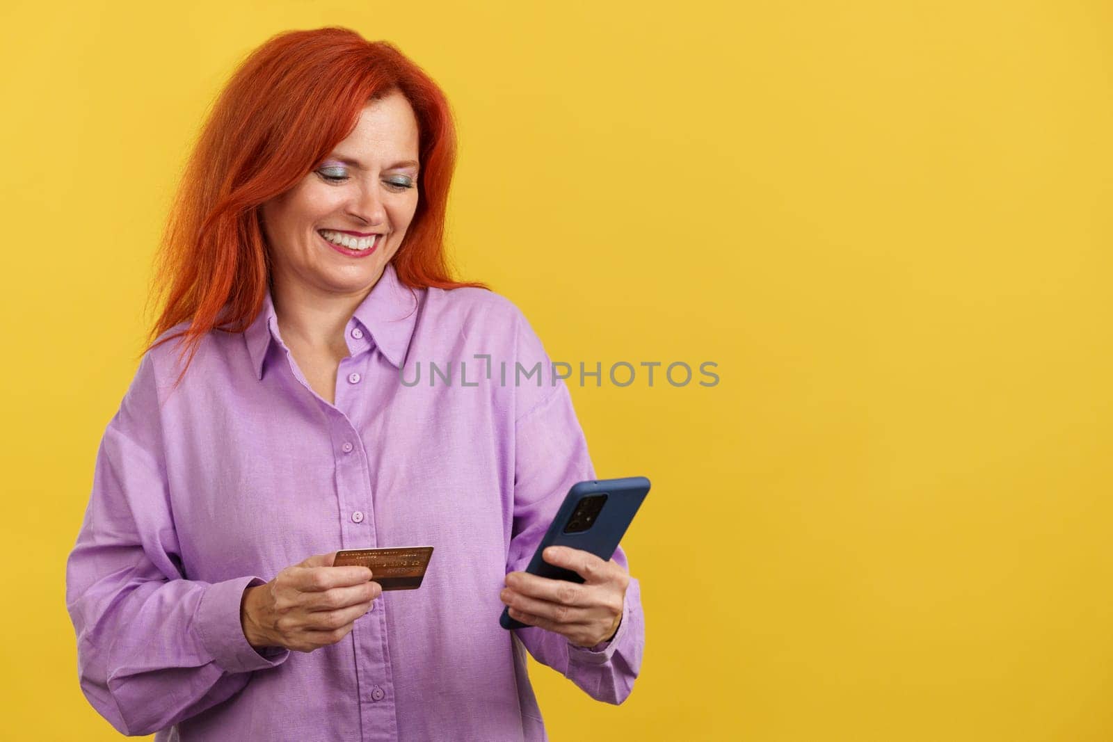 Happy mature redheaded woman using the mobile and card to shopping online in studio with yellow background