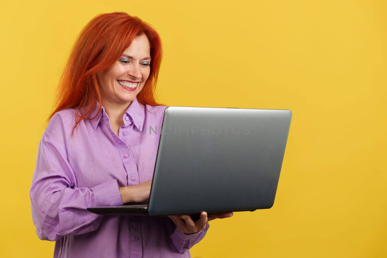 Redheaded mature woman smiling while using a laptop in studio with yellow background