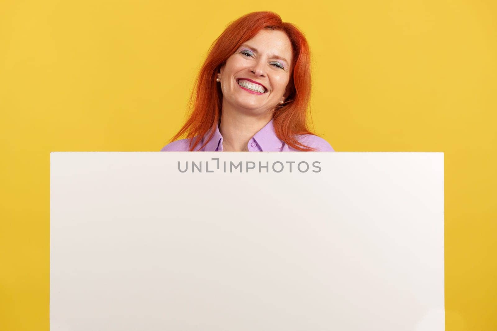 Mature redheaded woman holding a blank panel while smiling at the camera in studio with yellow background