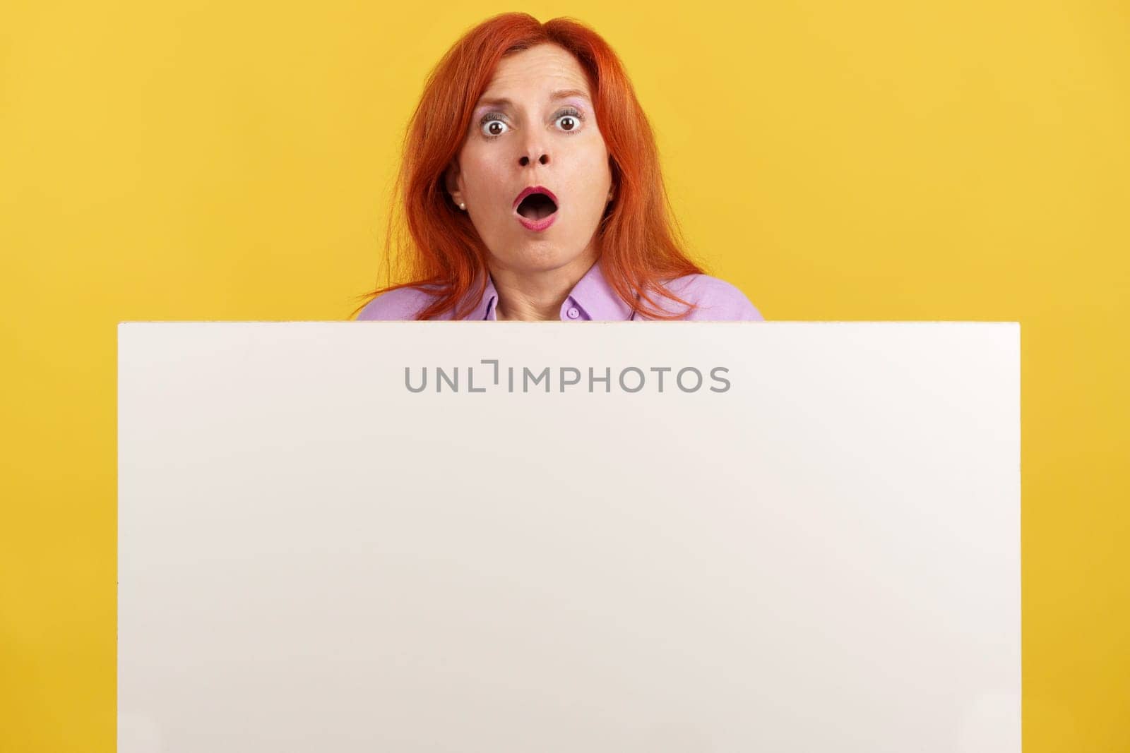 Amazed redheaded mature woman hair holding a blank panel and looking at camera with the mouth opened in studio with yellow background