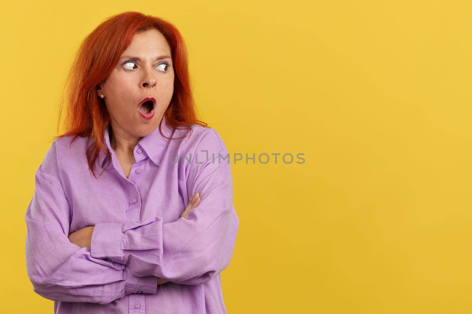 Surprised redheaded mature woman looking away with the mouth opened in studio with yellow background