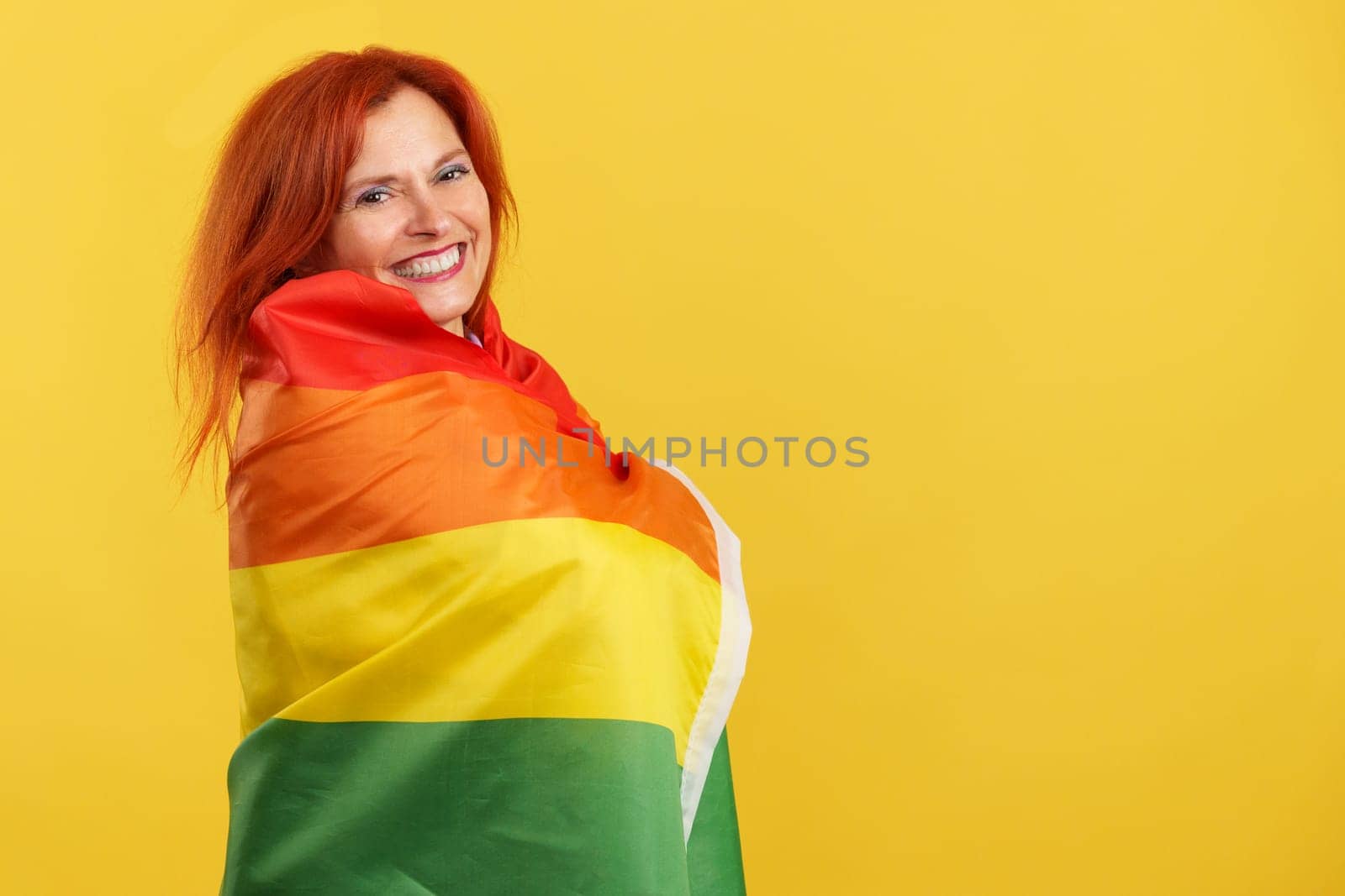 Happy redheaded mature woman wrapped with a lgbt rainbow flag in studio with yellow background