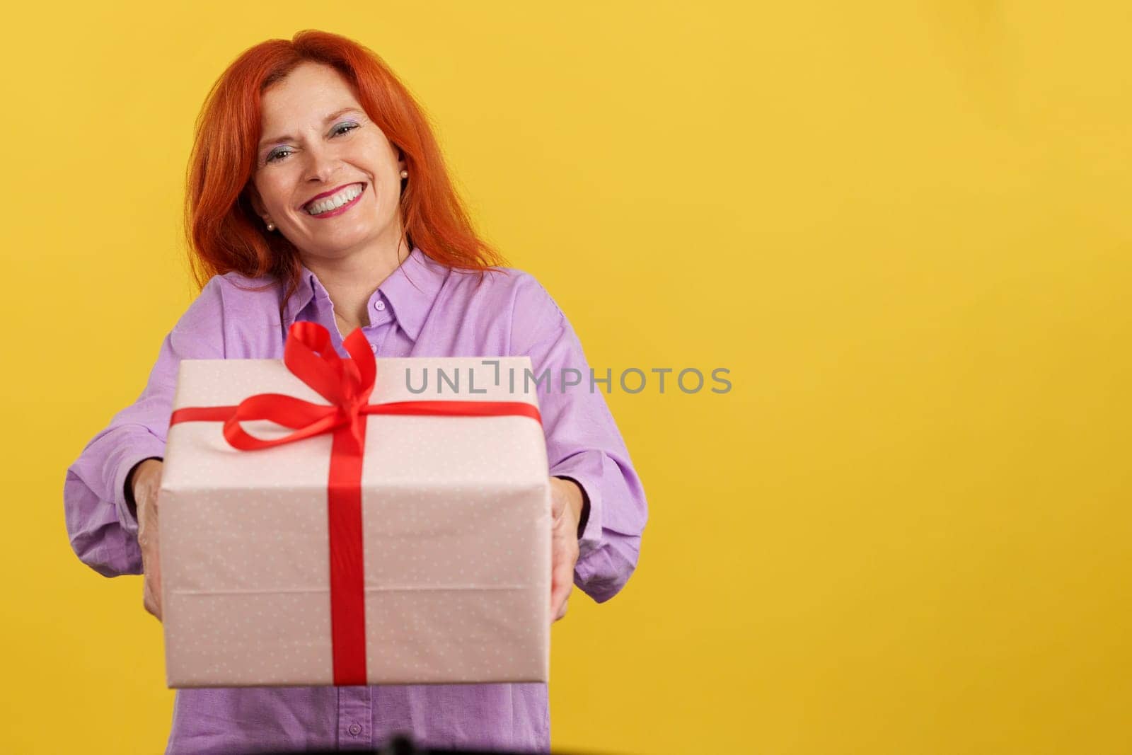 Mature woman with red-tinted hair delivering a gift for mother's day in studio with yellow background