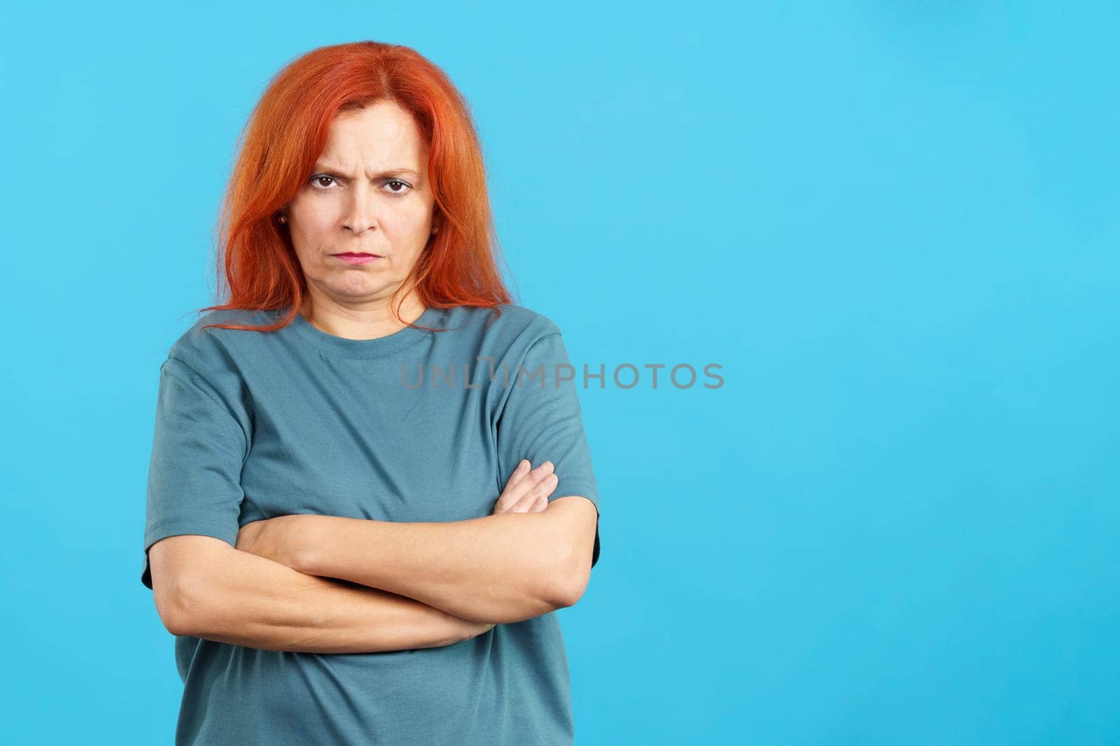 Mature redheaded woman crossing the arms with an angry expression in studio with blue background