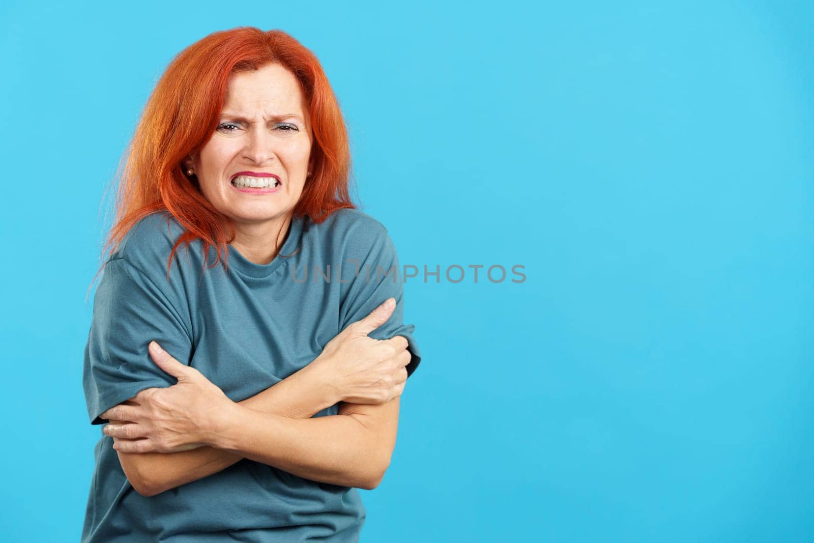 Mature redheaded woman looking at camera gesturing she is cold in studio with blue background