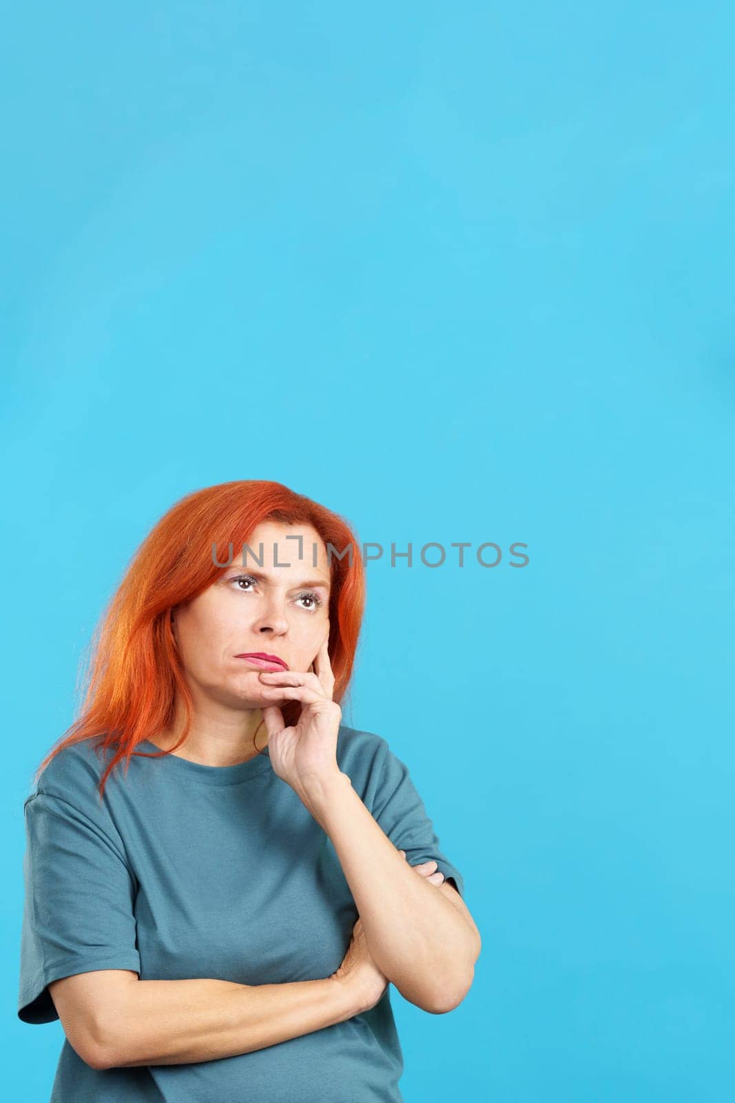 Bored redheaded mature woman standing and looking up in studio with blue background