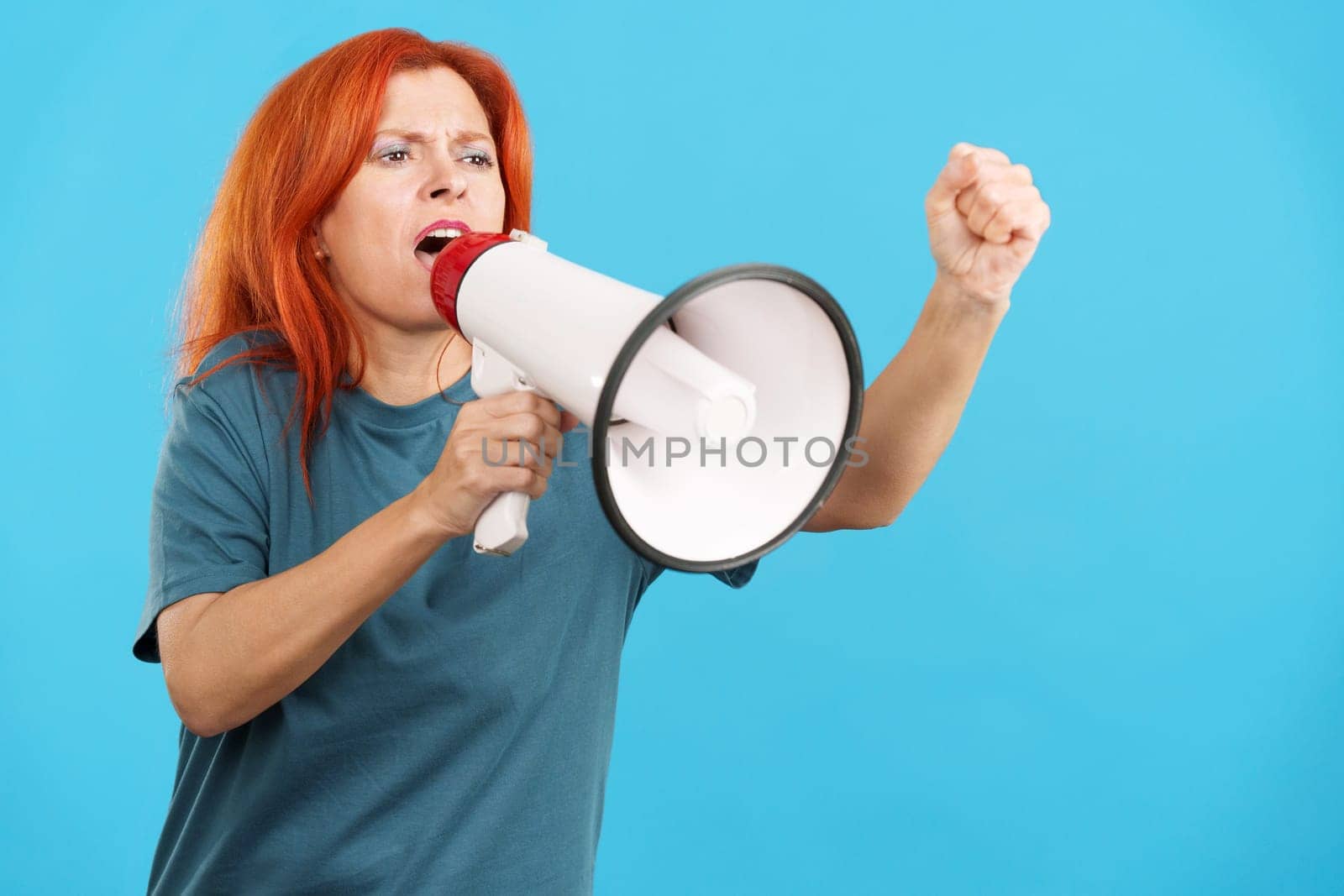 Upset redheaded mature woman yelling using a loudspeaker in studio with blue background