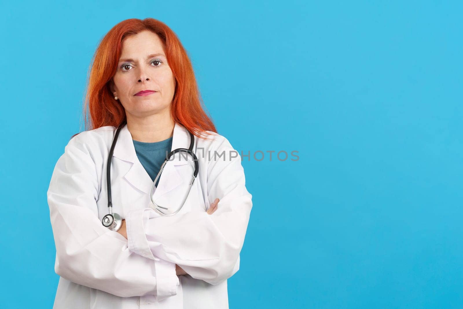 Serious mature redheaded female doctor standing with arms crossed in studio with blue background