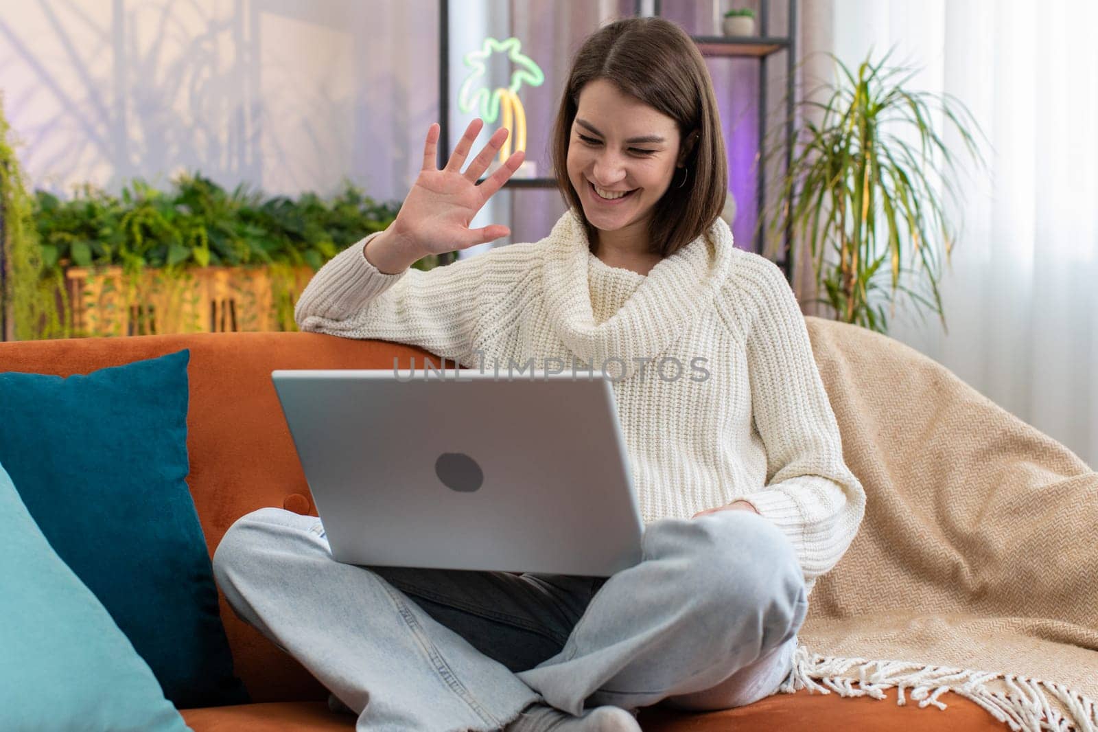 Young woman sitting on couch looking at camera, making video webcam conference call with friends or family, enjoying pleasant conversation. Portrait of pretty girl laughing waving hello at home room