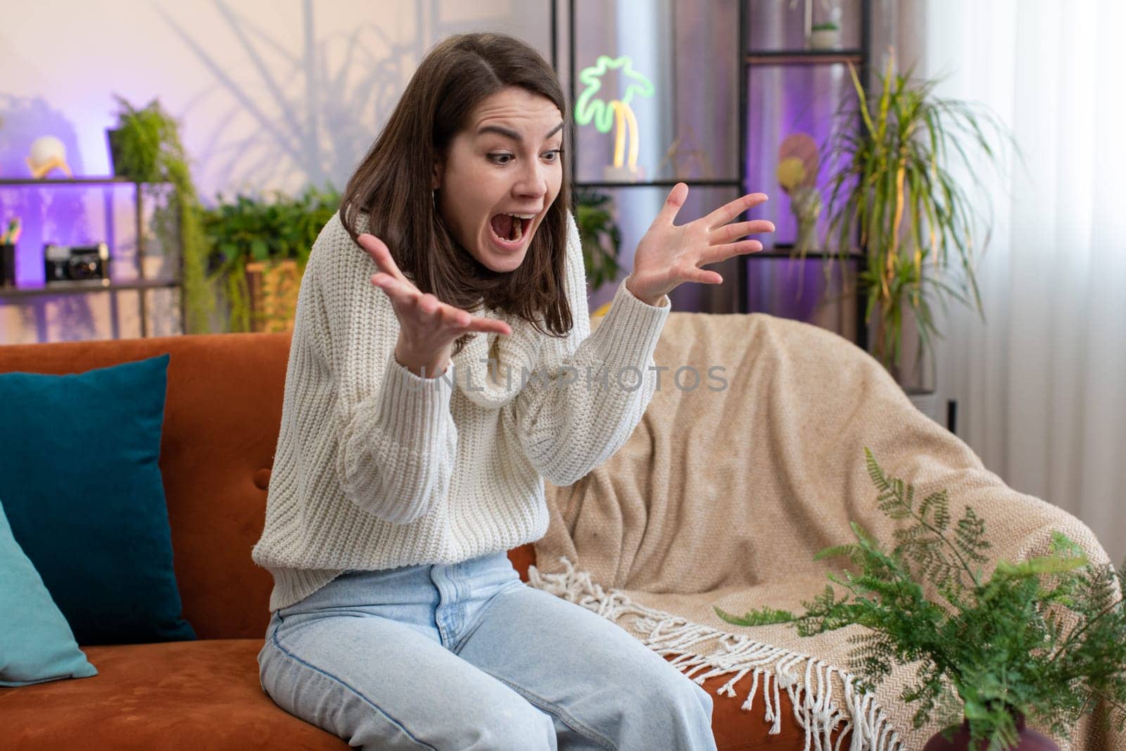 Stressed upset young woman screams from stress tension problems panic horror hopelessness fear surprise shock expresses gestures rage panic attack at home room apartment. Furious girl sitting on couch