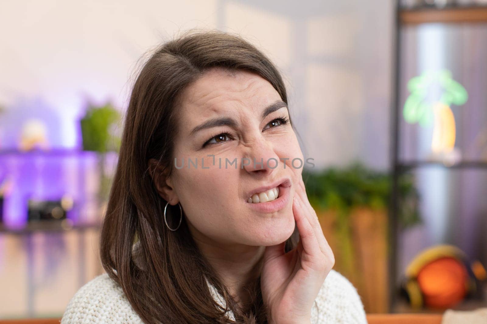 Dental problems. Portrait of young woman touching cheek, closing eyes with expression of terrible suffer from painful toothache, sensitive teeth, cavities. Girl sitting on couch at home room apartment