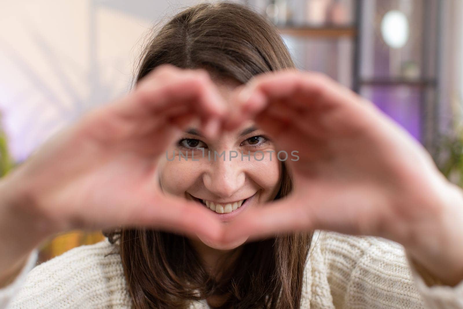 I love you. Happy young woman at home living room couch makes symbol of love, showing heart sign to camera, express romantic feelings express sincere positive feelings. Charity, gratitude, donation