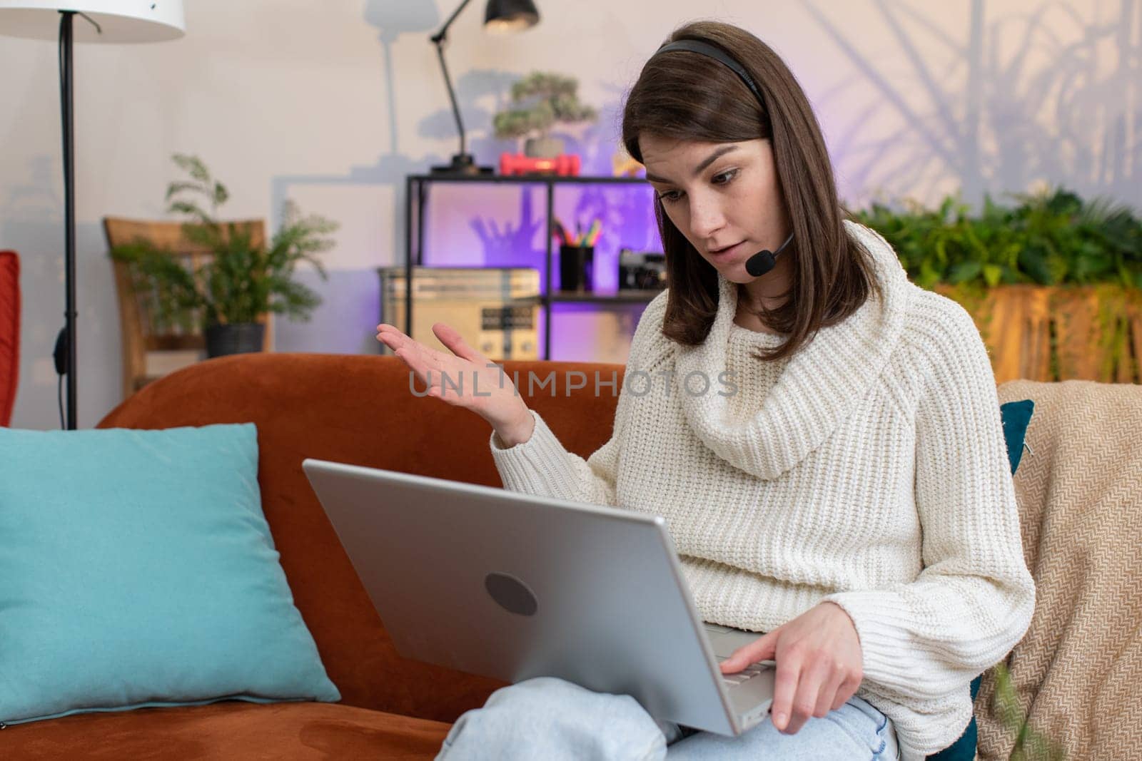Woman wearing headset, freelance worker, call center or support service operator helpline, having talk with client or colleague communication support waving hello at home room apartment. Girl on couch