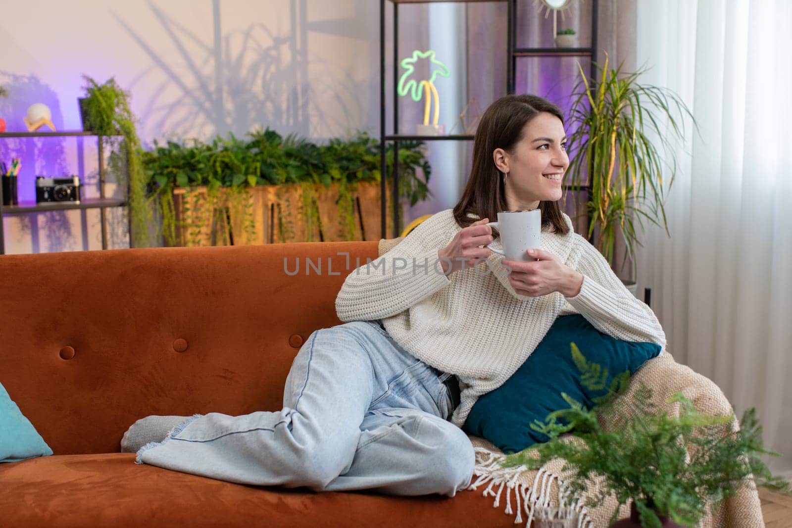 Portrait of attractive smiling young woman drinking a cup of warm coffee or herbal tea at home living room apartment in the morning. Pretty girl lying on sofa enjoying comfortable relaxing alone