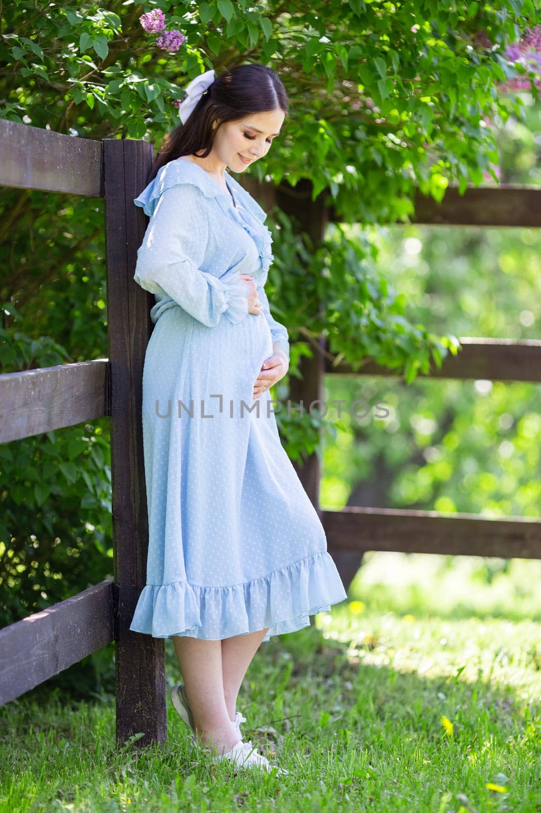 a smiling pregnant girl in a blue dress stands near a wooden fence by Zakharova