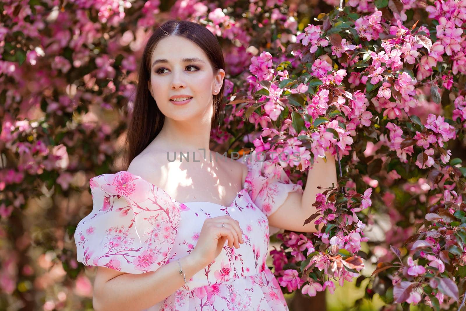 A charming smiling brunette in a pink dress standing near pink blooming apple trees, holding a blooming branch, in the spring in the garden, looks to the side. Copy space